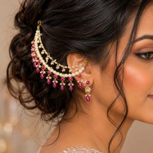 Puede incluir: Un conjunto de pendiente tipo ear cuff y accesorio para el cabello en tono dorado, con dos cadenas drapeadas adornadas con pequeñas perlas blancas y piedras magenta colgantes en forma de lágrima. Conecta la oreja con el cabello y lleva un pendiente.