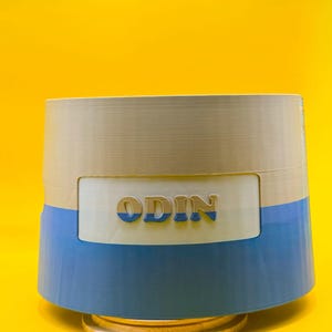 Pode incluir: Um recipiente cilíndrico com uma seção superior bege, uma seção inferior azul e uma faixa branca no meio. A palavra "ODIN" é exibida em uma fonte 3D na faixa branca. O recipiente está sobre uma base dourada em um fundo amarelo.