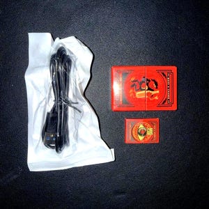 Puede incluir: Un cable de carga USB negro está envuelto en papel blanco. Dos cajas rojas de naipes están al lado del cable. Una caja es más grande que la otra. Las cajas tienen diseños dorados y negros.