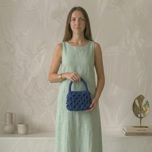 Peut inclure: Une robe sans manches en lin vert clair associée à un petit sac à main bleu profond doté d'un motif crocheté texturé en forme d'écailles superposées et d'une anse arrondie unique. Le sac est tenu au centre, révélant son motif complexe.