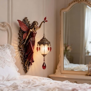 Op de afbeelding: Een wandlamp met een gebeeldhouwde feeënfiguur in een donkerrode jurk met bijpassende vleugels. De fee houdt een hangende lantaarn vast met een matglazen kap, versierd met kleine rode bladvormige bedels en een grote rode kristallen druppel.