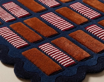 May include: A rectangular rug with a scalloped edge featuring a deep navy blue base. The surface is decorated with a repeating grid pattern of raised, rectangular tufted blocks in alternating solid rust orange and white and red striped textures.