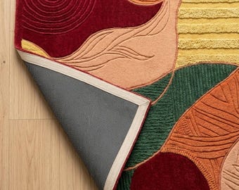 May include: A rectangular area rug featuring a tufted abstract pattern in deep red, mustard yellow, forest green, and peach. The rug has a dark gray non-slip backing visible where one corner is folded over, revealing a light beige fabric border.