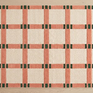 May include: A rectangular area rug featuring a grid pattern of peach-colored vertical and horizontal bands. The intersections of these bands are accented with small, dark green rectangular blocks, while the squares between are off-white.