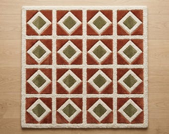 May include: A square rug featuring a repeating geometric pattern of sixteen squares arranged in a four-by-four grid. Each square contains a central olive green diamond surrounded by a thick white border, set against a rust orange background.