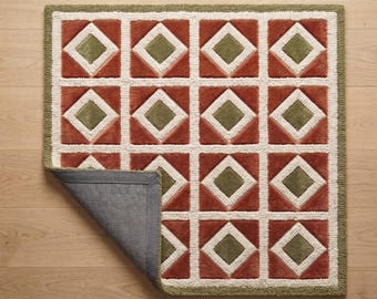 May include: A square tufted rug featuring a repeating geometric pattern of olive green diamonds centered within terracotta orange squares, outlined by cream-colored borders. The rug has a dark gray backing visible where one corner is folded over.