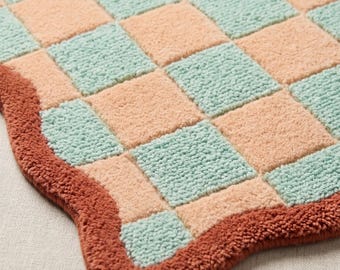 May include: A tufted rug featuring a repeating checkerboard pattern in soft peach and light mint green squares. The rug has a thick, textured pile and is finished with a solid, wavy-edged border in a deep, earthy terracotta brown color.