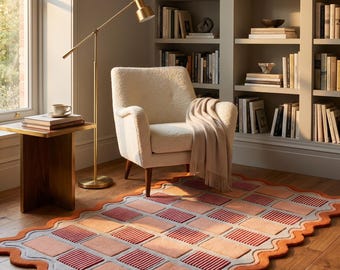 May include: A rectangular area rug featuring a wavy border and a grid pattern of alternating light pink and dark red striped rectangular blocks. The rug has a light blue base layer and a thick, textured orange outer edge, laid on a wooden floor.