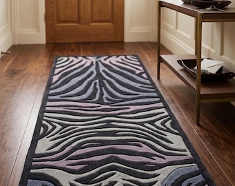 May include: A rectangular runner rug featuring a repeating zebra stripe pattern in shades of charcoal gray, light gray, and muted lavender. The rug has a thick, textured pile with a dark charcoal border framing the entire design on all four sides.