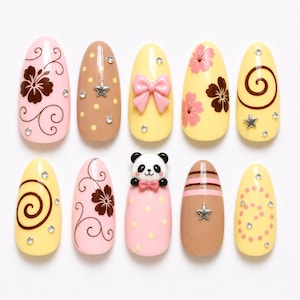 Può includere: Un set di dieci unghie finte a mandorla con vari design, tra cui motivi floreali marroni, fiocchi rosa, un viso di panda in 3D, spirali marroni, pois rosa e borchie a forma di stella argentata, il tutto arricchito da piccoli strass trasparenti.