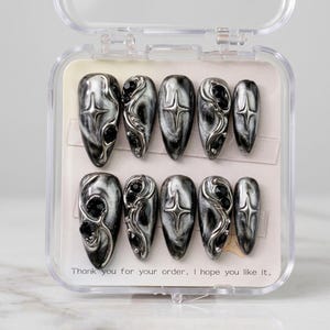May include: A set of ten press-on nails in a clear plastic case. The nails feature a black and silver marbled pattern with raised silver metallic accents, including star shapes and small black rhinestones. The text reads: Thank you for your order. I hope you like it.