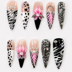 Puede incluir: Un conjunto de diez uñas postizas en forma de estilete con diseños variados que incluyen rayas de cebra en blanco y negro, manchas de leopardo, detalles florales rosas en 3D, remolinos plateados metálicos y pedrería incrustada.