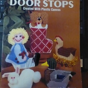 May include: A book cover titled "DOOR STOPS" showcasing needlework designs. The cover features plastic canvas creations: a dog in a stocking, a doll, a sheep, a duck, and a chicken. The book is by Kappie Originals Ltd.