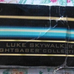 Op de afbeelding: Een gedeelte van een zwarte doos met gouden en blauwe strepen. De tekst "LUKE SKYWALKER LIGHTSABER COLLECTIBLE" wordt weergegeven in een gouden rechthoek.