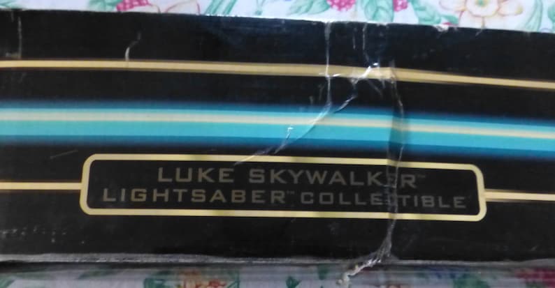 Op de afbeelding: Een gedeelte van een zwarte doos met gouden en blauwe strepen. De tekst "LUKE SKYWALKER LIGHTSABER COLLECTIBLE" wordt weergegeven in een gouden rechthoek.