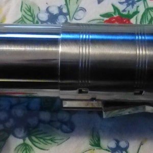 Op de afbeelding: Een metalen lichtzwaard prop met een zilveren en zwarte handgreep. Het lichtzwaard heeft een blauwe bedieningsknop en een gouden embleem. Het blad is wit en de handgreep is gedetailleerd met ribbels en groeven.