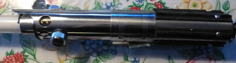 Op de afbeelding: Een metalen lichtzwaard prop met een zilveren en zwarte handgreep. Het lichtzwaard heeft een blauwe bedieningsknop en een gouden embleem. Het blad is wit en de handgreep is gedetailleerd met ribbels en groeven.