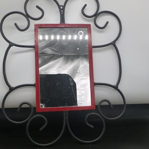 May include: A black metal wall mirror with a red frame. The mirror has a decorative scrolled design.