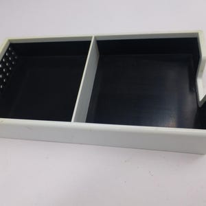 May include: A rectangular, two-compartment organizer with a white exterior and black interior. One side features a series of small, evenly spaced holes. The other side has a notched design.