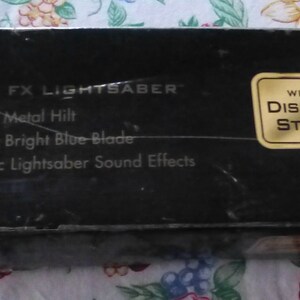 Op de afbeelding: Zwarte doos met de tekst "FORCE FX LIGHTSABER" en kenmerken: "Realistic Metal Hilt", "Glowing Bright Blue Blade" en "Authentic Lightsaber Sound Effects". Een gouden sticker leest "WITH DISPLAY STAND". De doos is voor een lichtzwaard.