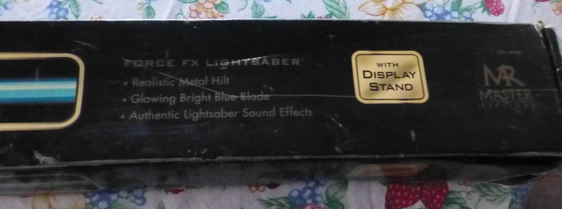 Op de afbeelding: Zwarte doos met de tekst "FORCE FX LIGHTSABER" en kenmerken: "Realistic Metal Hilt", "Glowing Bright Blue Blade" en "Authentic Lightsaber Sound Effects". Een gouden sticker leest "WITH DISPLAY STAND". De doos is voor een lichtzwaard.
