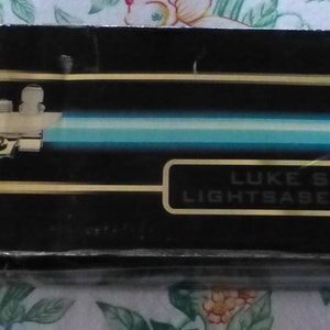 Op de afbeelding: Zwarte doos met een illustratie van een lichtzwaard en een blauw lemmet. Het lichtzwaard heeft een cr&egrave;mekleurig handvat. De tekst "LUKE SKYWALKER LIGHTSABER COLLECTIBLE" staat in goud op de doos.