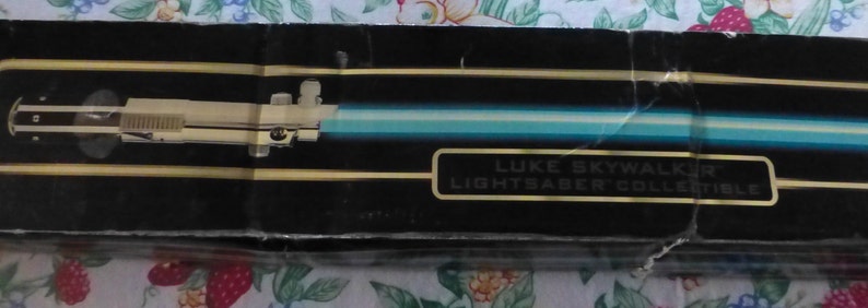 Op de afbeelding: Zwarte doos met een illustratie van een lichtzwaard en een blauw lemmet. Het lichtzwaard heeft een cr&egrave;mekleurig handvat. De tekst "LUKE SKYWALKER LIGHTSABER COLLECTIBLE" staat in goud op de doos.