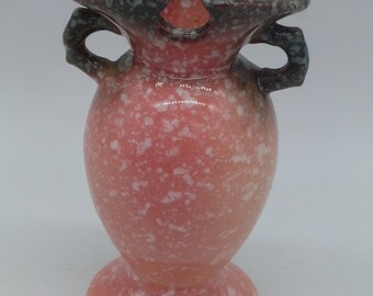 May include: A pink ceramic vase with white speckles and two handles. The vase has a scalloped top and a wide base.