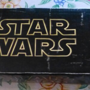 Op de afbeelding: Zwarte doos met de gouden tekst "STAR WARS" in een vetgedrukte, omlijnde lettertype. De doos lijkt vintage, met wat slijtage zichtbaar. Het Star Wars-logo is de belangrijkste focus.