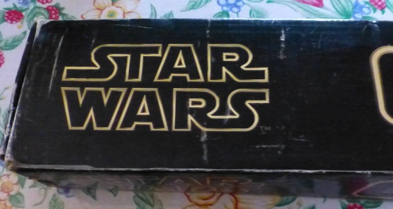 Op de afbeelding: Zwarte doos met de gouden tekst "STAR WARS" in een vetgedrukte, omlijnde lettertype. De doos lijkt vintage, met wat slijtage zichtbaar. Het Star Wars-logo is de belangrijkste focus.