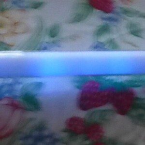 Op de afbeelding: Een lang, wit, cilindrisch object met een blauw gloeiend licht dat van binnenuit komt. Het licht lijkt gesegmenteerd, met afwisselende banden van helderder en donkerder blauw. Het object staat tegen een bloemenachtergrond.