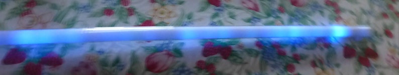 Op de afbeelding: Een lang, wit, cilindrisch object met een blauw gloeiend licht dat van binnenuit komt. Het licht lijkt gesegmenteerd, met afwisselende banden van helderder en donkerder blauw. Het object staat tegen een bloemenachtergrond.