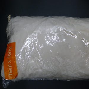 May include: A white twin x-long pillow in a clear plastic package. The pillow is rectangular and appears fluffy. An orange label with the text "twin x-long" is visible on the package.