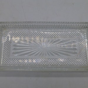 May include: A clear, rectangular glass dish with a textured surface. The dish features a central sunburst design and a patterned border. The edges are slightly rounded, and the overall design is simple and elegant.
