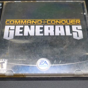 May include: A clear plastic CD case for the video game "Command & Conquer Generals." The game title is in large, silver block letters with a gold accent. The case has a "Teen" rating and the EA Games logo.