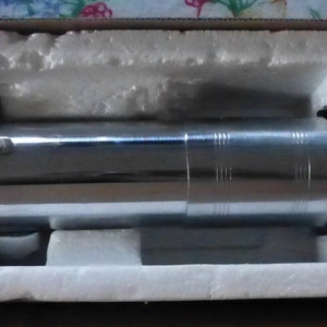 Op de afbeelding: Een zilverkleurig lichtzwaard rekwisiet, met zwarte accenten, rust in een beschermende, met schuim gevoerde doos. Het lichtzwaard heeft een gedetailleerde handgreep met diverse metalen componenten en een cilindrisch ontwerp. De algehele esthetiek suggereert een sciencefiction thema.