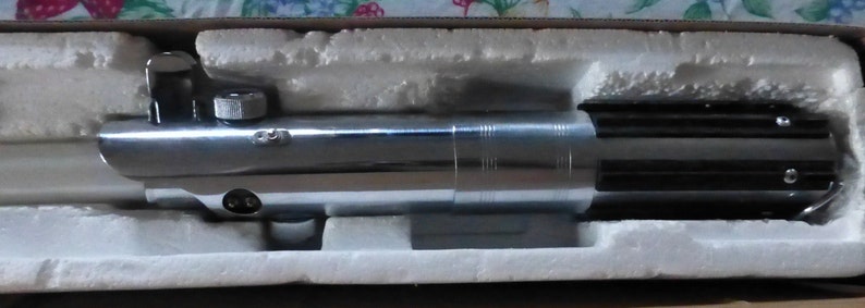 Op de afbeelding: Een zilverkleurig lichtzwaard rekwisiet, met zwarte accenten, rust in een beschermende, met schuim gevoerde doos. Het lichtzwaard heeft een gedetailleerde handgreep met diverse metalen componenten en een cilindrisch ontwerp. De algehele esthetiek suggereert een sciencefiction thema.