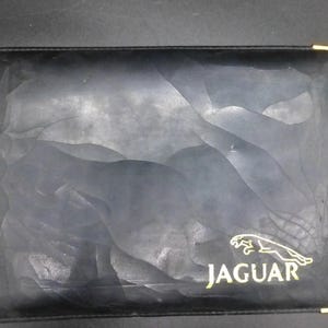 May include: Black leather-like wallet with a textured surface and gold-colored corner accents. The Jaguar logo and text are in gold. The wallet has a unique, abstract design.