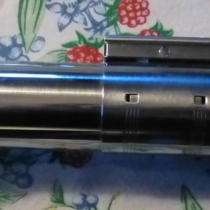 Op de afbeelding: Een metallic zilveren en zwarte lichtzwaard prop met een rode knop en een wit blad. De prop heeft een gedetailleerd ontwerp met verschillende metalen componenten en een rechthoekig bovenstuk. Het lichtzwaard is een replica van een populair sciencefictionwapen.