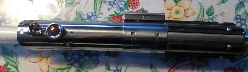 Op de afbeelding: Een metallic zilveren en zwarte lichtzwaard prop met een rode knop en een wit blad. De prop heeft een gedetailleerd ontwerp met verschillende metalen componenten en een rechthoekig bovenstuk. Het lichtzwaard is een replica van een populair sciencefictionwapen.