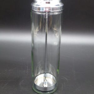 May include: A clear glass jar with a shiny, silver lid and base. A long, thin metal rod extends from the lid to the base. The jar is cylindrical and appears to be empty. The lid has a small, decorative knob.