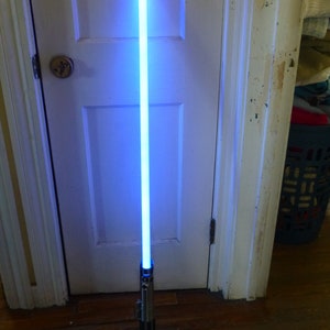 Op de afbeelding: Een blauw lichtzwaard met een zilveren en zwarte handgreep staat rechtop tegen een witte deur. Het blad straalt een helder, blauw licht uit. De handgreep heeft gedetailleerde metalen accenten.