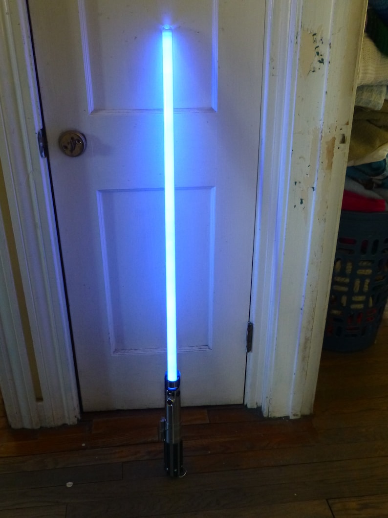 Op de afbeelding: Een blauw lichtzwaard met een zilveren en zwarte handgreep staat rechtop tegen een witte deur. Het blad straalt een helder, blauw licht uit. De handgreep heeft gedetailleerde metalen accenten.