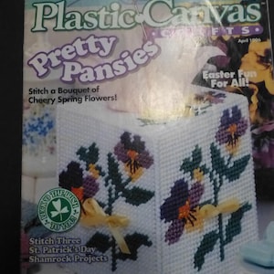 Op de afbeelding: Een witte plastic canvas tissuebox met geborduurde paarse, gele en groene viooltjes. De hoes is versierd met gestikte bloemen en een geel lint. De omslag van het tijdschrift luidt "Pretty Pansies" en "Stitch a Bouquet of Cheery Spring Flowers!"