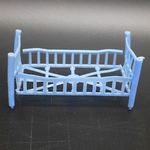 May include: A light blue miniature crib, featuring a slatted design for the sides and headboard. The crib has four vertical posts and a decorative center piece. The toy is made of plastic and is designed for dolls or other small toys.