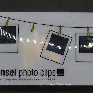 Puede incluir: Una tarjeta blanca con un gráfico de tres marcos de fotos colgados de una cuerda para tender la ropa con pinzas para la ropa. El texto "Tinsel photo clips" está impreso en negro en la parte inferior de la tarjeta. El texto "Pinces photo - Clips para fotos - Bilderklammern - フォトクリップ - 相片夹" está impreso en negro debajo del texto principal.