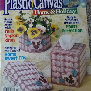 May include: A magazine cover titled "Plastic Canvas Home & Holiday" with projects for the home. Featured are needlepoint projects including a tissue box, napkin rings, and a decorative box, all adorned with pansy flower designs. The magazine is dated March-April 2000.