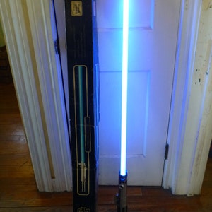 Op de afbeelding: Een blauw lichtzwaard met een zilveren handvat is verlicht, naast de zwarte doos. De doos heeft het Star Wars-logo en een diagram van het lichtzwaard. Het lichtzwaard straalt een helder, effen blauw licht uit.