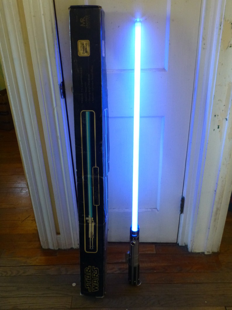 Op de afbeelding: Een blauw lichtzwaard met een zilveren handvat is verlicht, naast de zwarte doos. De doos heeft het Star Wars-logo en een diagram van het lichtzwaard. Het lichtzwaard straalt een helder, effen blauw licht uit.