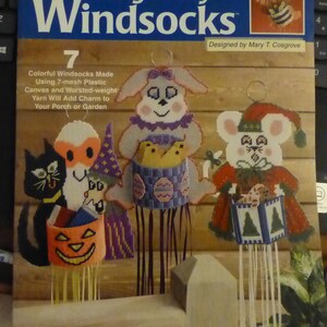 May include: A craft book cover with the title "Sitting Pretty Windsocks" and a picture of seven colorful windsocks. The windsocks are made with plastic canvas and worsted weight yarn. The book is designed by Mary T. Cosgrove.